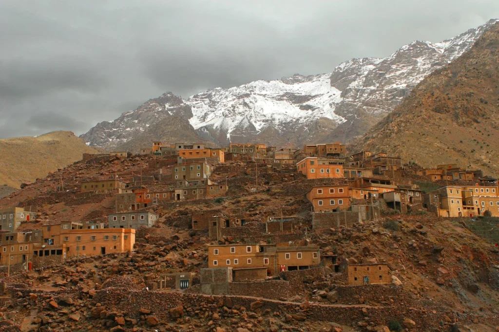 An Amazigh village of earth and stone houses on a slope below snow-capped High Atlas peaks